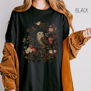 May include: Black t-shirt featuring a detailed illustration of an owl perched on a flowering branch. The owl has brown and white feathers, and the flowers are pink and white. The word "BLACK" is printed in white on the upper right.