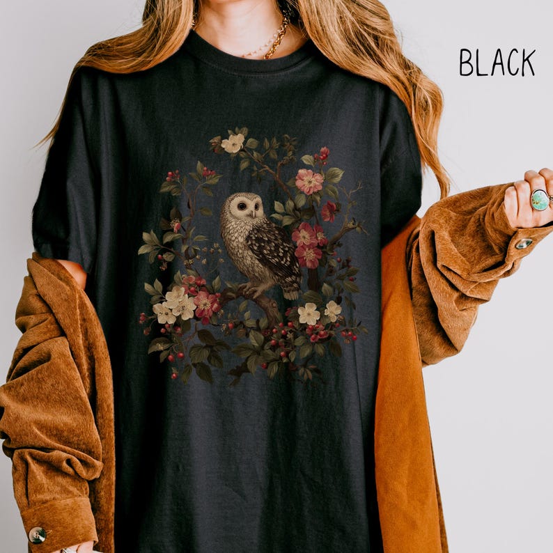 May include: Black t-shirt featuring a detailed illustration of an owl perched on a flowering branch. The owl has brown and white feathers, and the flowers are pink and white. The word "BLACK" is printed in white on the upper right.