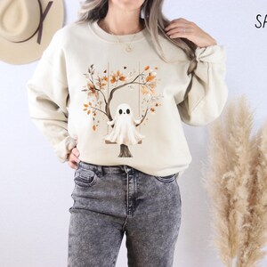 May include: Sand-colored sweatshirt featuring a whimsical ghost on a swing, set against a tree with autumn leaves. The design is printed on a long-sleeved, crew-neck top. The word "SAND" is printed in black on the upper right.