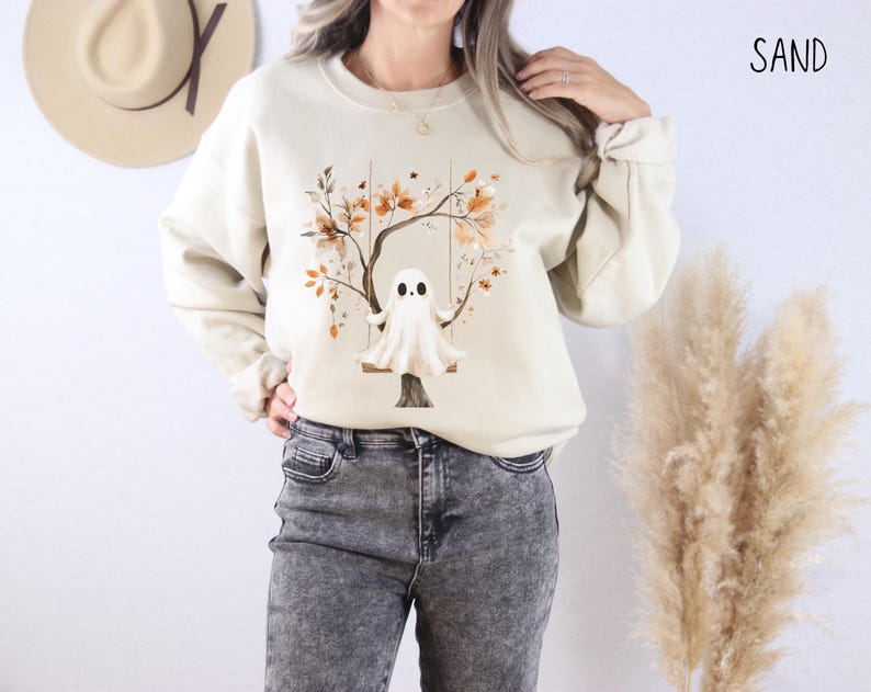 May include: Sand-colored sweatshirt featuring a whimsical ghost on a swing, set against a tree with autumn leaves. The design is printed on a long-sleeved, crew-neck top. The word "SAND" is printed in black on the upper right.