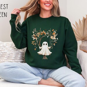 May include: Forest green sweatshirt featuring a whimsical ghost on a swing, surrounded by autumn-colored leaves and branches. The text "FOREST GREEN" is printed in black at the top left. The sweatshirt is a crew neck style.