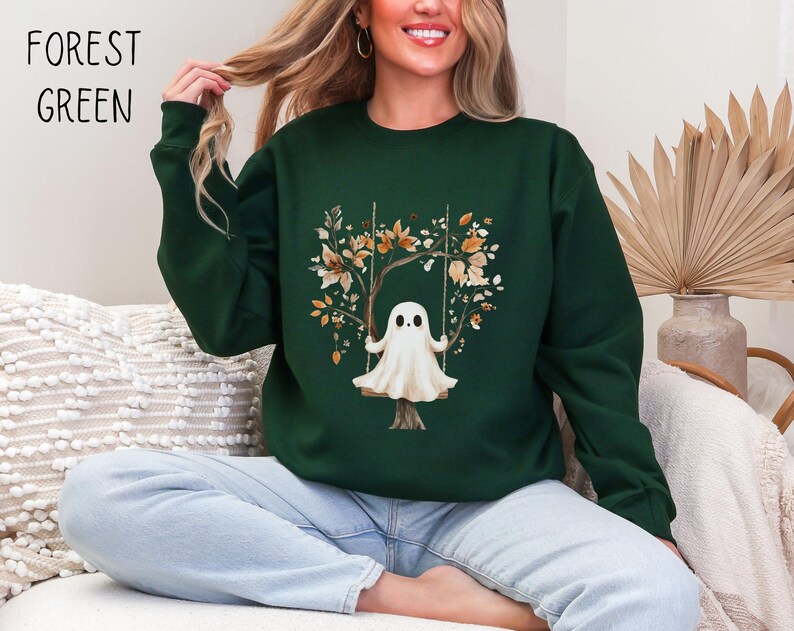 May include: Forest green sweatshirt featuring a whimsical ghost on a swing, surrounded by autumn-colored leaves and branches. The text "FOREST GREEN" is printed in black at the top left. The sweatshirt is a crew neck style.
