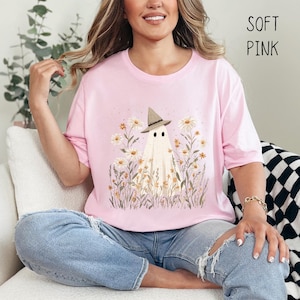 K&ouml;nnte beinhalten: Hellrosa T-Shirt mit einem Geistermotiv, der einen Hexenhut tr&auml;gt, umgeben von G&auml;nsebl&uuml;mchen und hohem Gras. Der Text "SOFT PINK" ist oben rechts aufgedruckt. Die Person tr&auml;gt zerrissene blaue Jeans.