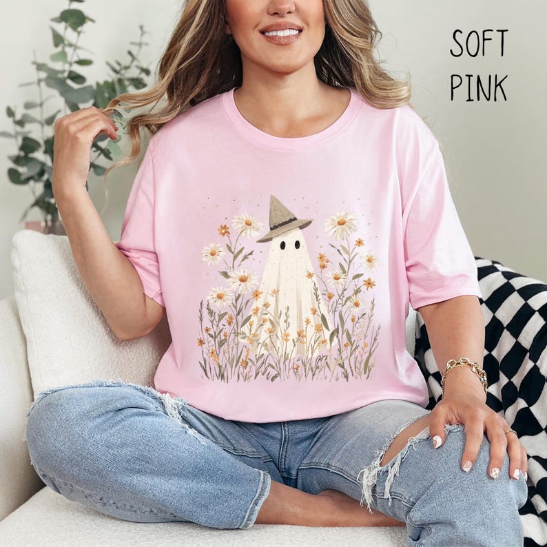 K&ouml;nnte beinhalten: Hellrosa T-Shirt mit einem Geistermotiv, der einen Hexenhut tr&auml;gt, umgeben von G&auml;nsebl&uuml;mchen und hohem Gras. Der Text "SOFT PINK" ist oben rechts aufgedruckt. Die Person tr&auml;gt zerrissene blaue Jeans.