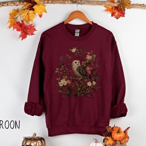 Op de afbeelding: Bordeaux sweatshirt met een gedetailleerde uil illustratie omringd door bloemen. De top met lange mouwen heeft een ronde hals en opgerolde manchetten. Het woord "MAROON" staat in het zwart linksonder.
