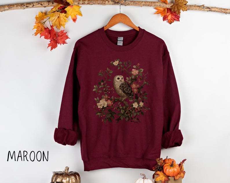 Op de afbeelding: Bordeaux sweatshirt met een gedetailleerde uil illustratie omringd door bloemen. De top met lange mouwen heeft een ronde hals en opgerolde manchetten. Het woord "MAROON" staat in het zwart linksonder.