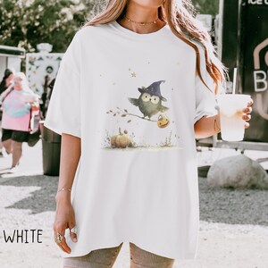 May include: White t-shirt featuring a watercolor illustration of a green owl wearing a witch hat, perched on a branch with a pumpkin and a jack-o'-lantern. The word "WHITE" is printed at the bottom left. A casual, oversized fit.