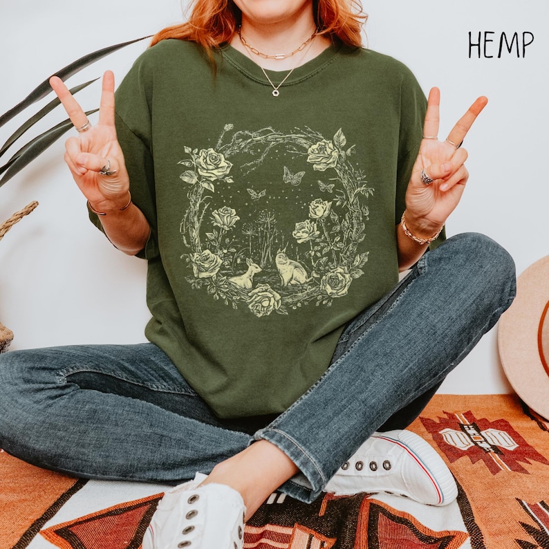 May include: Olive green sweatshirt with a circular floral design featuring roses, butterflies, a deer, and a cat. The word "HEMP" is printed in white on the upper right. The model is wearing blue jeans and white sneakers.