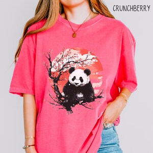 K&ouml;nnte beinhalten: Ein pinkfarbenes T-Shirt mit einem Aufdruck eines Pandas, der in einem Kirschbl&uuml;tenbaum vor einer roten Sonne sitzt.