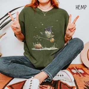 May include: Olive green sweatshirt featuring a whimsical Halloween owl wearing a witch hat, perched on a branch with a pumpkin. The word "HEMP" is printed in white on the upper right. The owl is holding a jack-o'-lantern. The sweatshirt is paired with blue jeans.