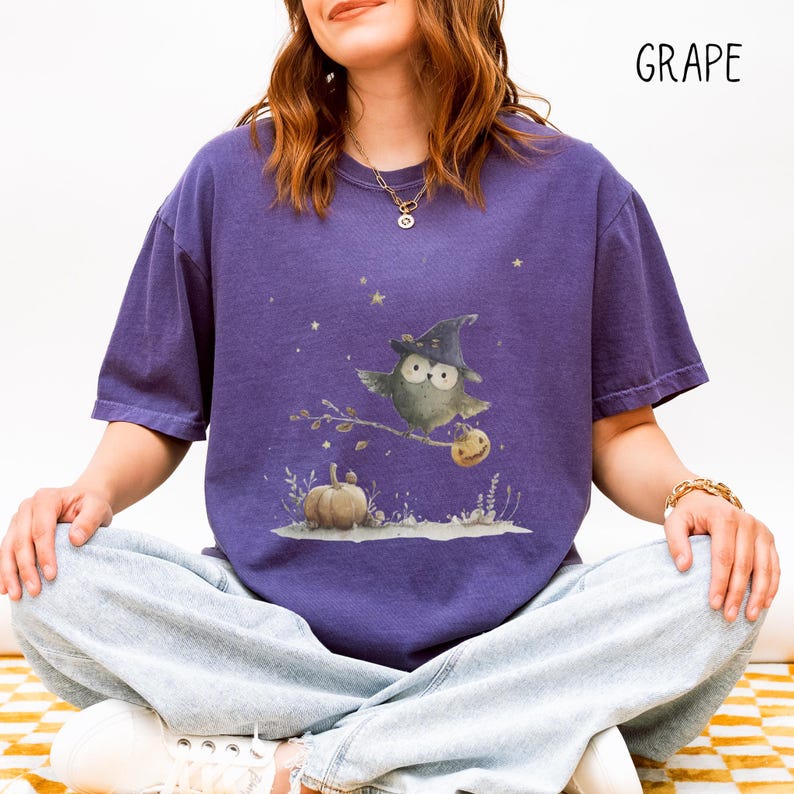 May include: A purple t-shirt featuring a whimsical Halloween design. The graphic depicts an owl wearing a witch hat, perched on a branch with a pumpkin. The word "GRAPE" is printed in the upper right corner.