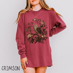 May include: Crimson long-sleeve shirt featuring a detailed illustration of an owl perched on a flowering branch. The owl is depicted with intricate feather patterns and is surrounded by delicate flowers and leaves. The word "CRIMSON" is printed at the bottom.