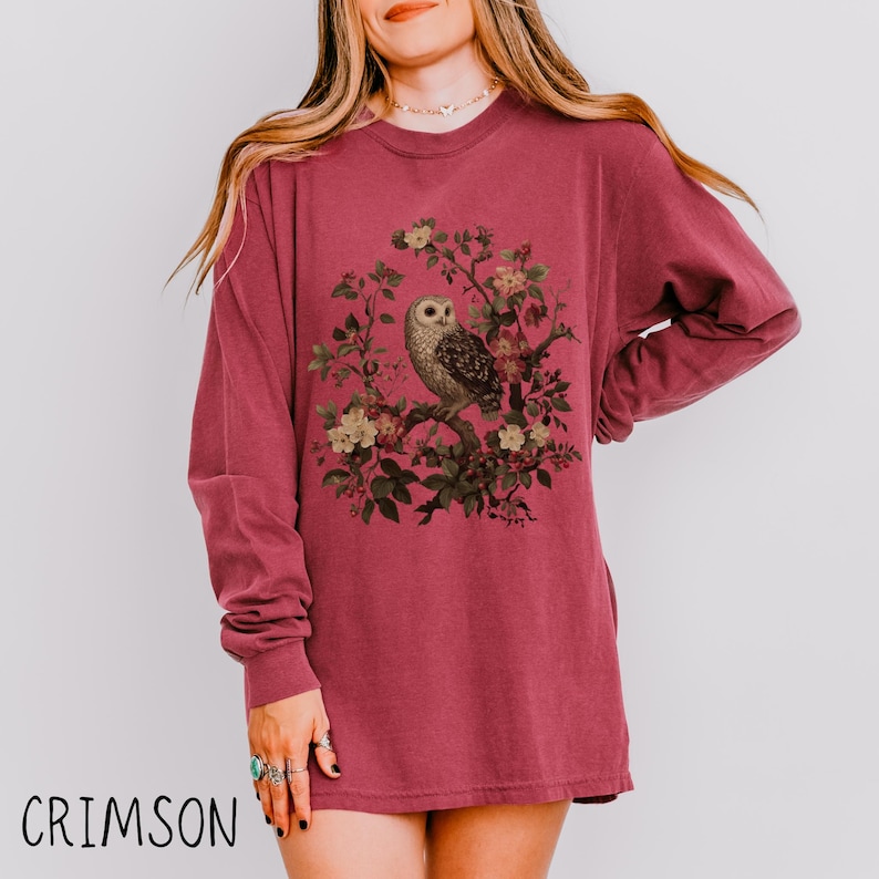 May include: Crimson long-sleeve shirt featuring a detailed illustration of an owl perched on a flowering branch. The owl is depicted with intricate feather patterns and is surrounded by delicate flowers and leaves. The word "CRIMSON" is printed at the bottom.