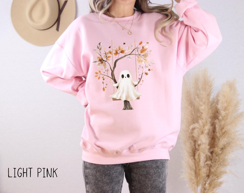 May include: Light pink sweatshirt featuring a whimsical illustration of a ghost on a swing beneath a tree with autumn leaves. The text "LIGHT PINK" is printed at the bottom. The sweatshirt is a comfortable, casual style.