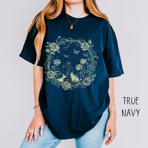 May include: Navy blue t-shirt featuring a floral wreath design with roses, butterflies, and two small animals. The graphic is in a light yellow color. The text "TRUE NAVY" is printed on the right side of the shirt.