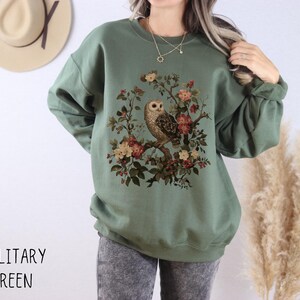Op de afbeelding: Militairgroen sweatshirt met een uil en bloemenontwerp. De illustratie toont een uil op een tak omringd door roze, rode en gele bloemen. De tekst "MILITARY GREEN" staat onderaan.