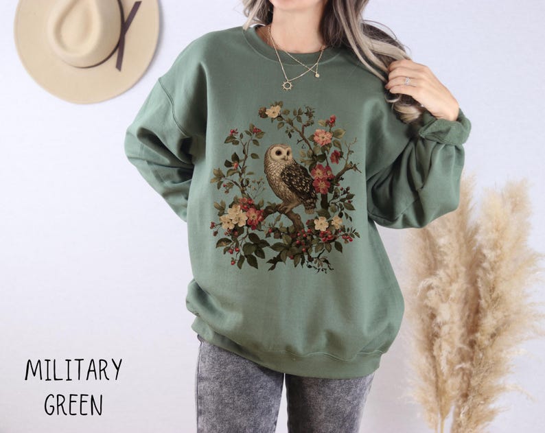 Op de afbeelding: Militairgroen sweatshirt met een uil en bloemenontwerp. De illustratie toont een uil op een tak omringd door roze, rode en gele bloemen. De tekst "MILITARY GREEN" staat onderaan.