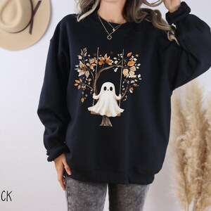 May include: Black sweatshirt featuring a whimsical illustration of a ghost on a swing set. The design includes a tree with autumn-colored leaves. The word "BLACK" is printed at the bottom. A cozy and stylish garment for Halloween or fall.