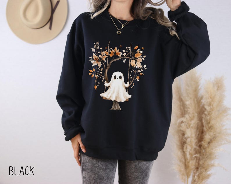 May include: Black sweatshirt featuring a whimsical illustration of a ghost on a swing set. The design includes a tree with autumn-colored leaves. The word "BLACK" is printed at the bottom. A cozy and stylish garment for Halloween or fall.