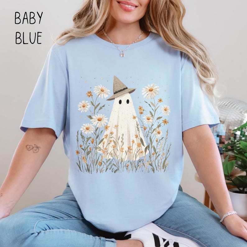 K&ouml;nnte beinhalten: Hellblaues T-Shirt mit einem Geist mit Hut, umgeben von G&auml;nsebl&uuml;mchen und Wildblumen. Der Text "BABY BLUE" ist oben links in Schwarz aufgedruckt. Ein charmantes Design f&uuml;r ein l&auml;ssiges T-Shirt.