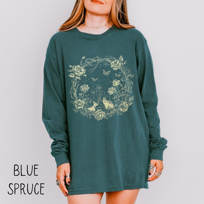 May include: Blue spruce long-sleeve shirt with a floral and animal graphic. The design features a wreath of roses, butterflies, and woodland creatures. The text "BLUE SPRUCE" is printed at the bottom of the shirt.