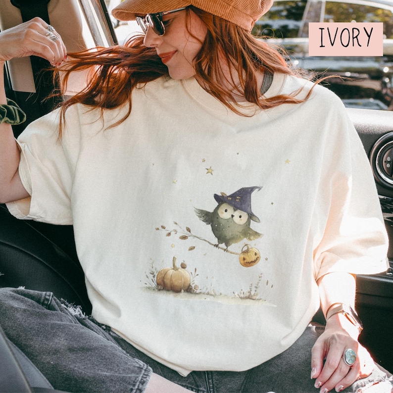 May include: Ivory-colored t-shirt featuring a watercolor illustration of a whimsical owl wearing a witch hat, perched on a branch with a pumpkin and jack-o'-lantern. The shirt has a relaxed fit and is perfect for Halloween or fall.
