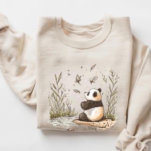 Könnte beinhalten: Ein beiger Pullover mit einem Aufdruck eines Pandas, der auf einem Stein sitzt, umgeben von grünen Pflanzen und Schmetterlingen. Der Panda schaut zu den Schmetterlingen auf.
