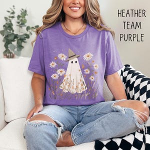 K&ouml;nnte beinhalten: Heather-lilafarbenes T-Shirt mit einem Geist, der einen Hexenhut tr&auml;gt, umgeben von G&auml;nsebl&uuml;mchen und Wildblumen. Der Text "HEATHER TEAM PURPLE" ist auf der rechten Seite aufgedruckt. Ein bequemes, l&auml;ssiges T-Shirt f&uuml;r den Alltag.