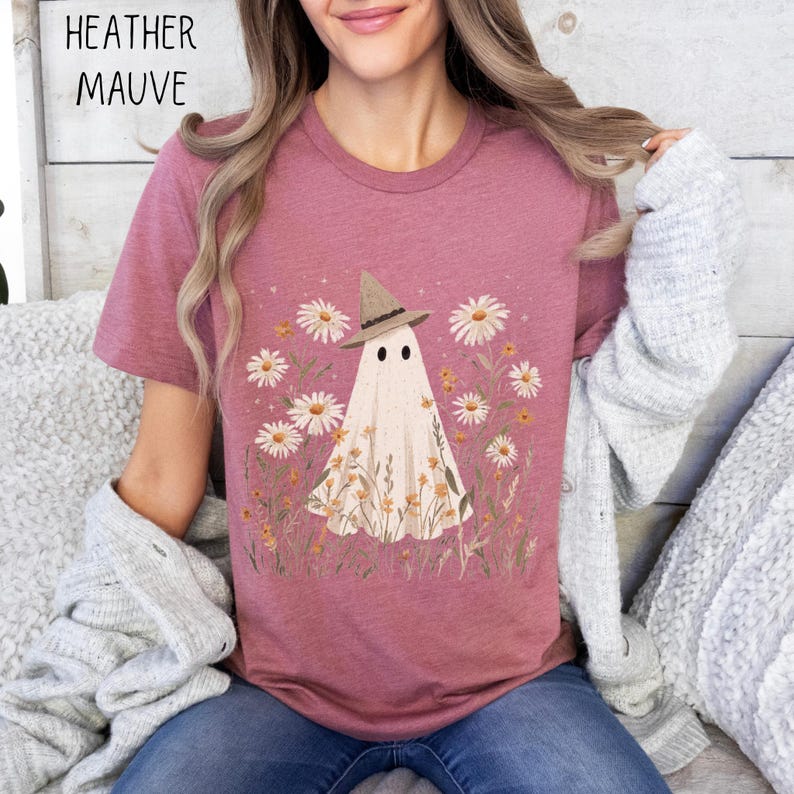 K&ouml;nnte beinhalten: Heather-Mauve-T-Shirt mit einem skurrilen Geist, der einen Hexenhut tr&auml;gt, umgeben von G&auml;nsebl&uuml;mchen und Wildblumen. Der Geist hat zwei schwarze Punktaugen. Der Text "HEATHER MAUVE" befindet sich in der oberen linken Ecke.