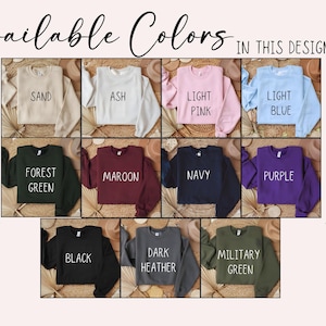Op de afbeelding: Afbeelding met een verscheidenheid aan sweatshirtkleuren. De tekst "Beschikbare kleuren in dit ontwerp" staat bovenaan. Kleuren zijn zand, as, lichtroze, lichtblauw, bosgroen, kastanjebruin, marineblauw, paars, zwart, donkergrijs en legergroen.