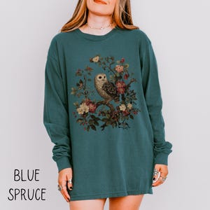 May include: Blue spruce long-sleeve shirt featuring a detailed illustration of an owl perched on a flowering branch. The owl is surrounded by colorful flowers and foliage. The words "BLUE SPRUCE" are printed at the bottom of the shirt.