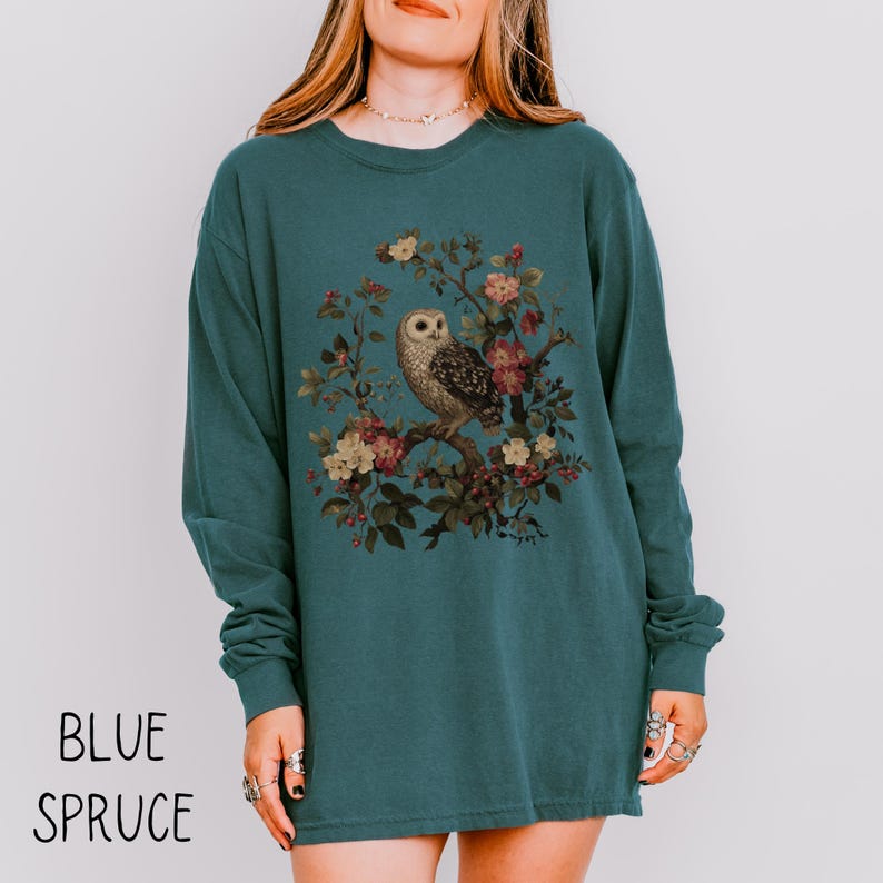 May include: Blue spruce long-sleeve shirt featuring a detailed illustration of an owl perched on a flowering branch. The owl is surrounded by colorful flowers and foliage. The words "BLUE SPRUCE" are printed at the bottom of the shirt.