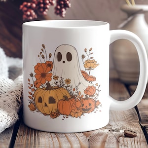 May include: White ceramic mug featuring a Halloween-themed design. The design includes a ghost, pumpkins, and orange flowers. The mug has a large handle and is set on a wooden surface.