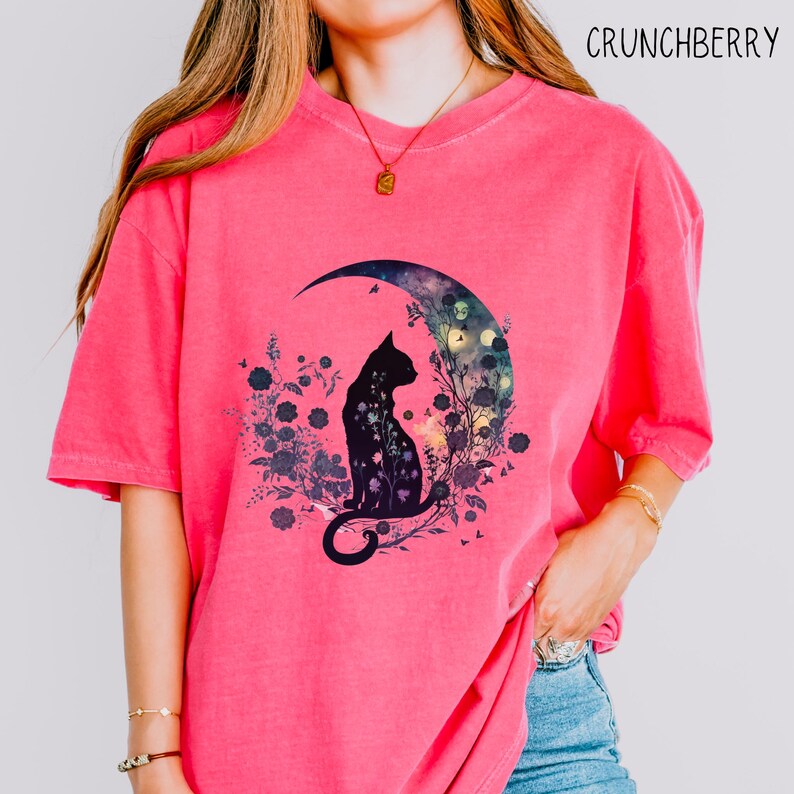 May include: A pink t-shirt featuring a black cat silhouette sitting on a crescent moon. The moon is filled with a floral design and a starry night sky. The text "CRUNCHBERRY" is at the top right.