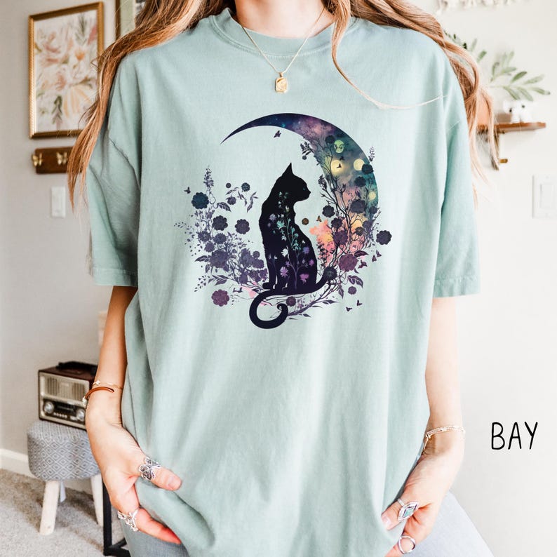 May include: A light green t-shirt featuring a black cat silhouette sitting inside a crescent moon. The moon is filled with colorful floral and botanical elements. The shirt has a relaxed fit.