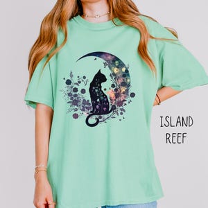May include: A light green t-shirt featuring a black cat silhouette sitting on a crescent moon with a floral design. The text "ISLAND REEF" is printed in black on the right side of the shirt. The shirt has short sleeves and a crew neck.
