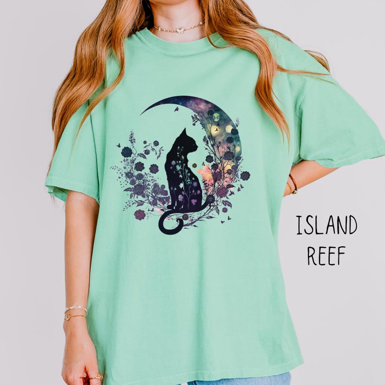 May include: A light green t-shirt featuring a black cat silhouette sitting on a crescent moon with a floral design. The text "ISLAND REEF" is printed in black on the right side of the shirt. The shirt has short sleeves and a crew neck.