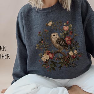 Op de afbeelding: Donkergrijs sweatshirt met een afbeelding van een uil op een bloeiende tak. De uil is bruin en wit, omringd door roze en gele bloemen. De tekst "DARK HEATHER" staat aan de linkerkant van de trui.