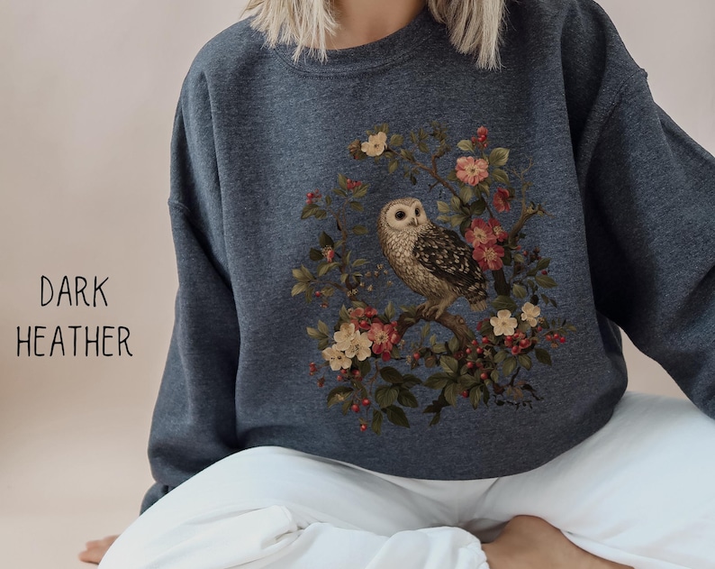 Op de afbeelding: Donkergrijs sweatshirt met een afbeelding van een uil op een bloeiende tak. De uil is bruin en wit, omringd door roze en gele bloemen. De tekst "DARK HEATHER" staat aan de linkerkant van de trui.