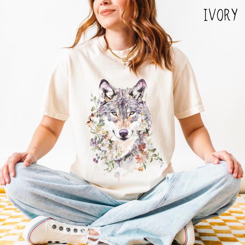 An ivory t-shirt featuring a detailed watercolor-style illustration of a gray wolf's face, framed by an arrangement of delicate wildflowers in muted tones of purple, orange, cream, and green. The floral elements blend into the fur, giving the design a soft, ethereal look. The woman wearing the shirt is seated cross-legged on a yellow and white checkered rug, with light-wash jeans and white sneakers, in a brightly lit, minimal setting.