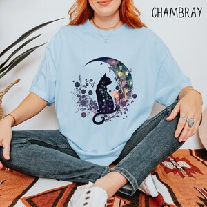 May include: Light blue t-shirt with a cat silhouette design. The cat is sitting on a crescent moon with a floral and galaxy-themed background. The word "CHAMBRAY" is printed in the upper right corner.