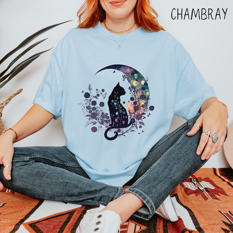 May include: Light blue t-shirt with a cat silhouette design. The cat is sitting on a crescent moon with a floral and galaxy-themed background. The word "CHAMBRAY" is printed in the upper right corner.