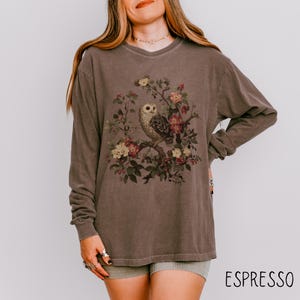 May include: Espresso-colored long-sleeve shirt featuring a vintage-style owl illustration perched on a flowering branch. The owl is detailed with brown and white feathers, surrounded by pink and yellow flowers. The word "ESPRESSO" is printed in white.