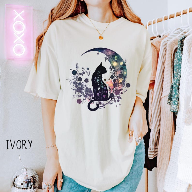 May include: Ivory-colored t-shirt featuring a black cat silhouette sitting on a crescent moon, surrounded by floral and celestial elements. The design includes a color palette of blues, purples, and greens. The word "IVORY" is printed on the bottom left.
