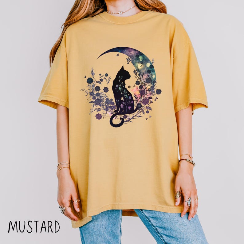May include: Mustard-colored t-shirt featuring a black cat silhouette seated on a crescent moon with a floral design. The moon has a galaxy-like pattern. The shirt's text reads "MUSTARD".