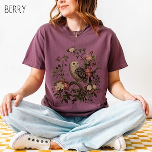 May include: Berry-colored t-shirt featuring a detailed illustration of an owl perched on a flowering branch. The owl has brown and white feathers, surrounded by pink and white blossoms. The word "BERRY" is printed in white in the upper left corner.