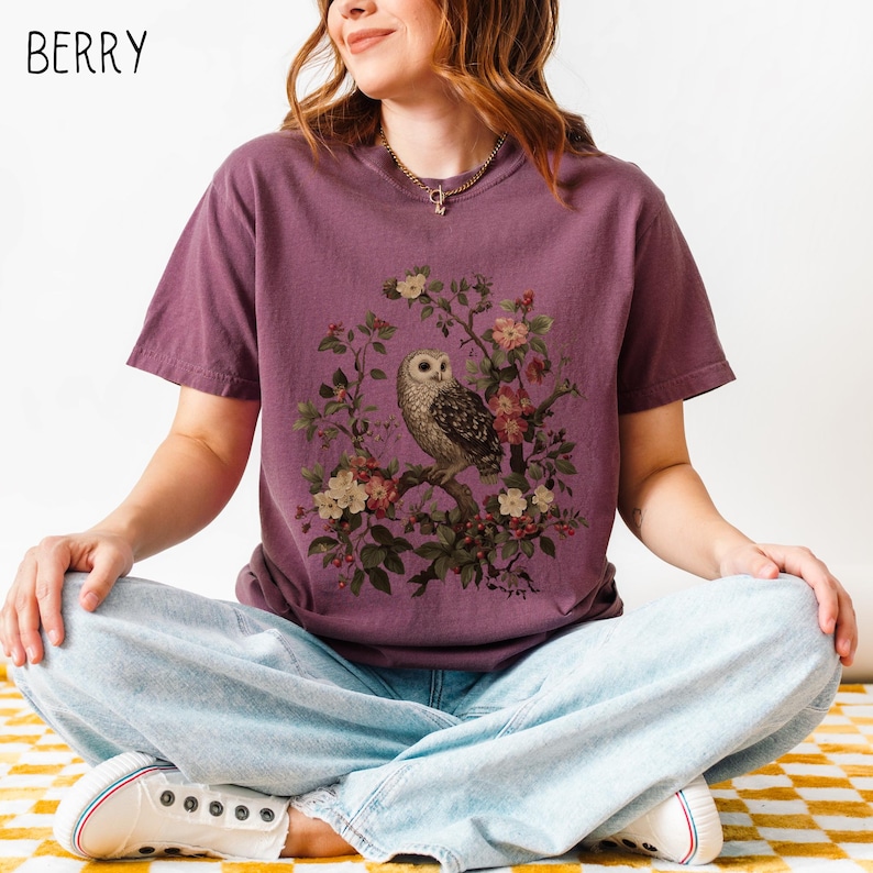May include: Berry-colored t-shirt featuring a detailed illustration of an owl perched on a flowering branch. The owl has brown and white feathers, surrounded by pink and white blossoms. The word "BERRY" is printed in white in the upper left corner.