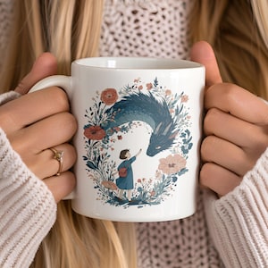 May include: White ceramic mug featuring a whimsical illustration of a blue dragon interacting with a small figure, surrounded by floral elements in shades of red, pink, and green. The mug is held by a person wearing a cream-colored sweater.