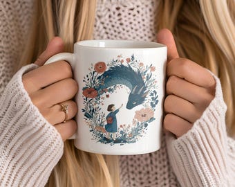 May include: White ceramic mug featuring a whimsical illustration of a blue dragon interacting with a small figure, surrounded by floral elements in shades of red, pink, and green. The mug is held by a person wearing a cream-colored sweater.