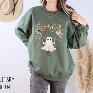 May include: Military green sweatshirt featuring a whimsical illustration of a ghost on a swing, set against a tree with autumn-colored leaves. The text "MILITARY GREEN" is printed at the bottom. The sweatshirt has long sleeves and a crew neck.